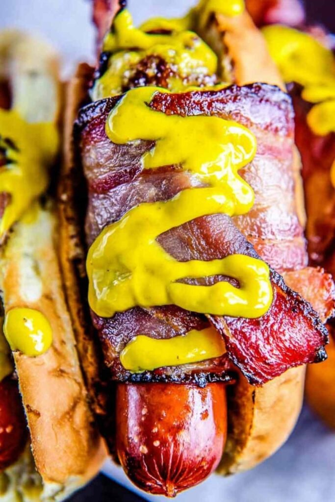 Bacon wrapped hot dogs grilling over low heat with crispy golden bacon on an outdoor grill