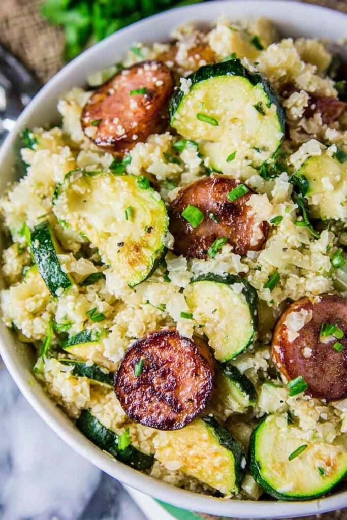 Cauliflower rice skillet with zucchini and kielbasa topped with melted cheddar cheese in a wide skillet
