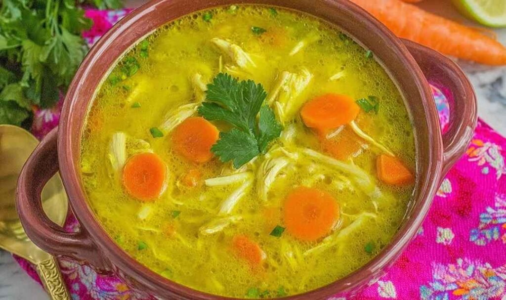 A bowl of creamy mulligatawny soup topped with avocado slices, fresh cilantro, and lime wedges on a wooden table