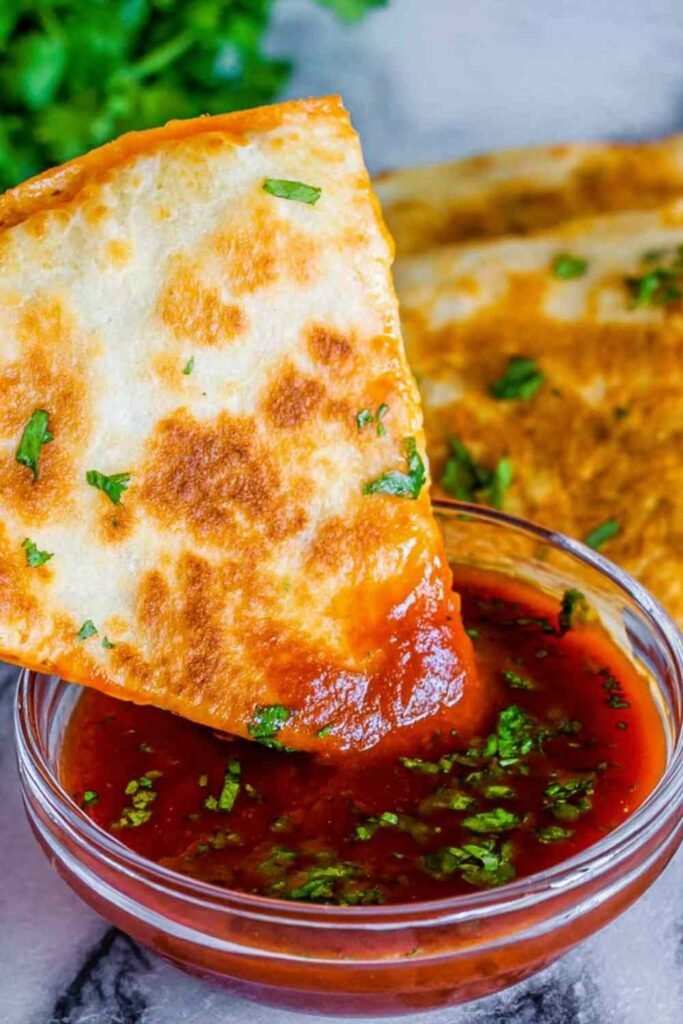 Crispy enchilada chicken quesadillas cut into wedges on a white plate served with red enchilada dipping sauce and fresh sliced avocado