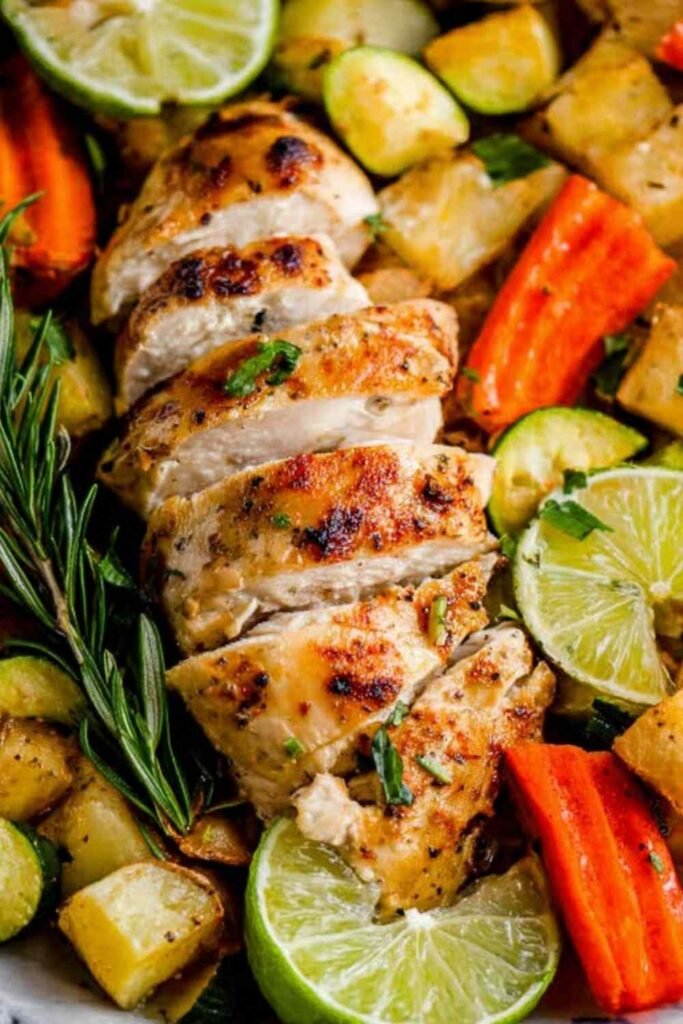 Raw sheet pan rosemary lime chicken thighs arranged with potatoes, carrots, zucchini, and lime wedges ready to roast