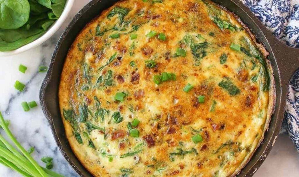 Golden spinach bacon frittata in a cast iron skillet topped with green onions and melted gouda cheese